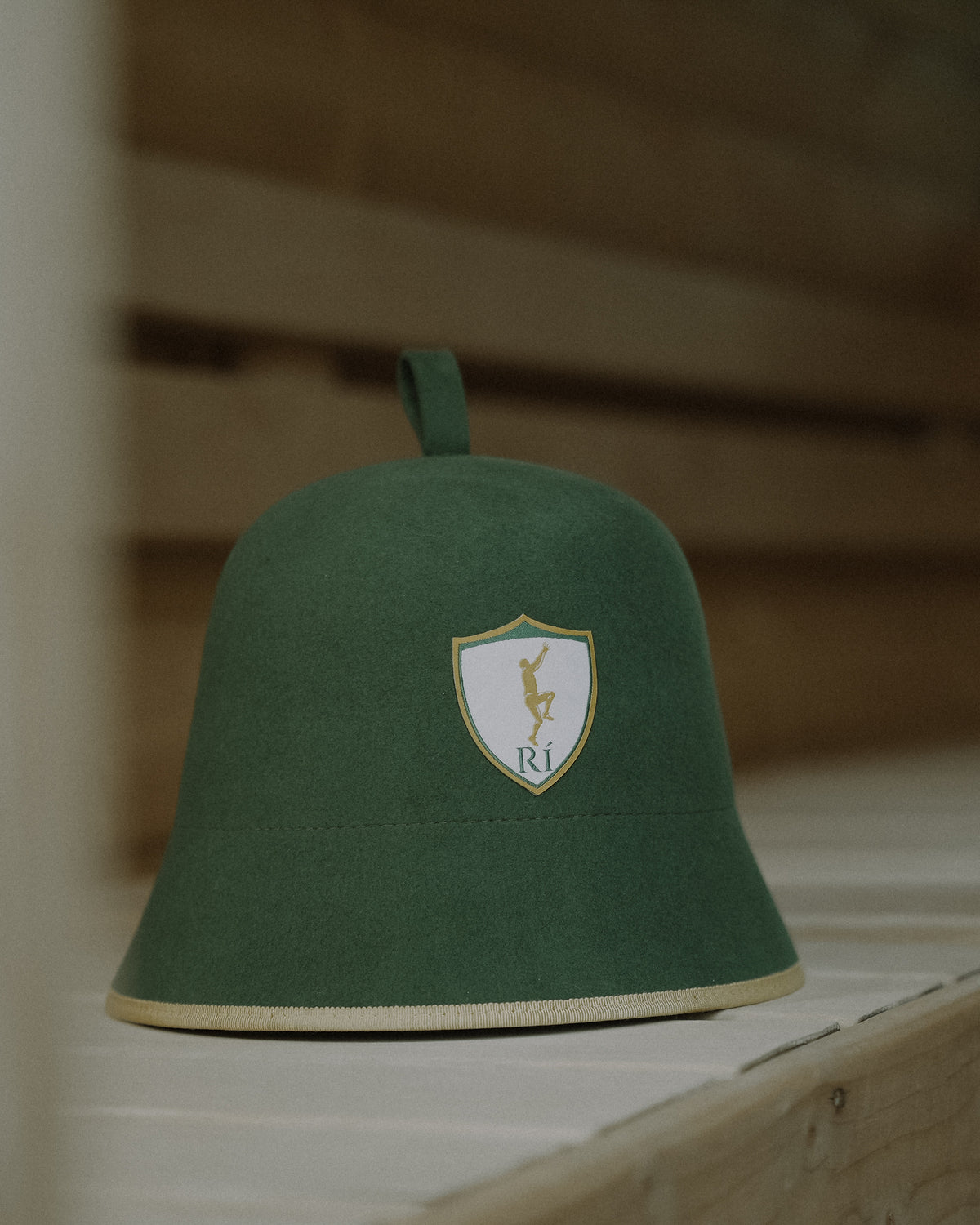 Rí Sauna Crown, deep green merino wool sauna hat on a sauna bench, shield crest facing forward